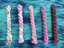 Load image into Gallery viewer, Five variegated mini skeins of yarn in shades of dusty pinks and purples lined up vertically on a background of variegated blue-green yarn