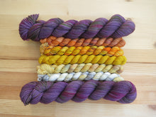 Load image into Gallery viewer, Five variegated mini skeins of yarn in shades of orange, yellows and cream flanked by two full sized variegated dusty purple skeins lined up horizontally on a pale wooden background