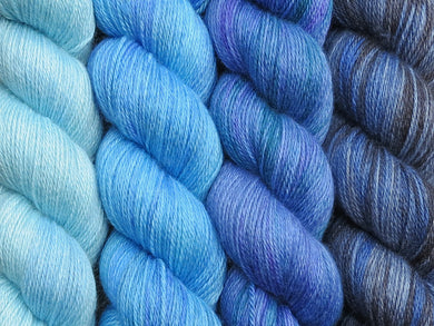 A close up of four skeins of yarn in various shades of blues from pale baby blue on the left through to a variegated black and blue on the right (Fhloston Geogradient Yarn Kit on Blue Chip Stocking)