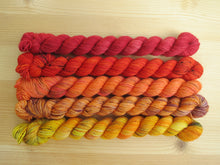 Load image into Gallery viewer, Five variegated mini skeins of yarn in shades of red, orange and yellows lined up horizontally on a pale wooden background