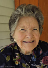 Load image into Gallery viewer, A photo by Megan Kenny of Rhelma Kenny (nee O'Reilly), taken in 2016. An older woman with greying hair is smiling at the camera. She wears a deep blue shirt with pink and white flowers with green leaves