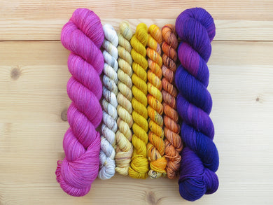 Five  mini skeins of yarn in shades of cream, yellow and orange flanked by two full sized skeins of bright pink on the left and deep purple on the right lined up vertically on a pale wooden background