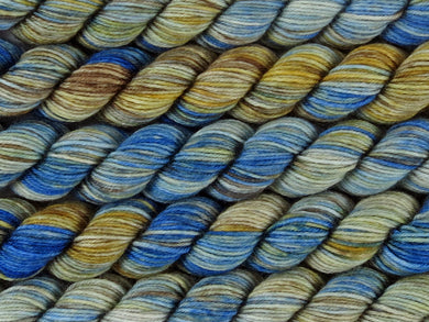 A close up of variegated blue, ecru, milk chocolate and golden yellow mini skeins of superwash merino and nylon 4ply fingering sock yarn arranged horizontally (The Gap on Tough Stocking Mini)