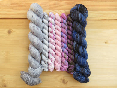 Five mini skeins of yarn in shades of white, pink and purple flanked by two full sized skeins of grey with purple speckles on the left and variegated blue black on the right lined up vertically on a pale wooden background