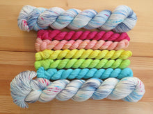 Load image into Gallery viewer, Five light bright rainbow coloured mini skeins of yarn flanked by two full sized variegated white, sky blue and grey with pink speckles skeins lined up horizontally on a pale wooden background
