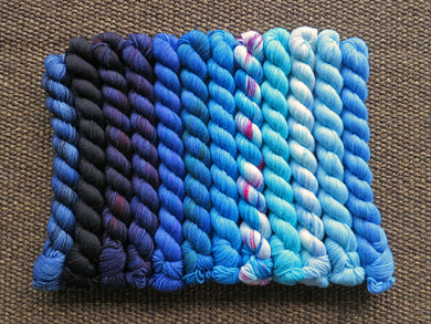 Twelve mini skeins of yarn in various shades of blue ranging from dark to light lined up on a brown woven background