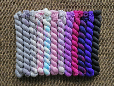 Twelve mini skeins of yarn in various colours ranging from greys through pinks to purples lined up on a brown woven background