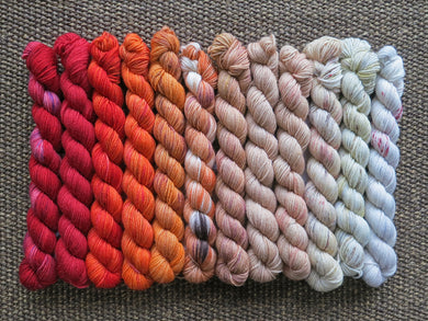 Twelve mini skeins of yarn in various colours ranging from reds through oranges to tan and beige lined up on a brown woven background