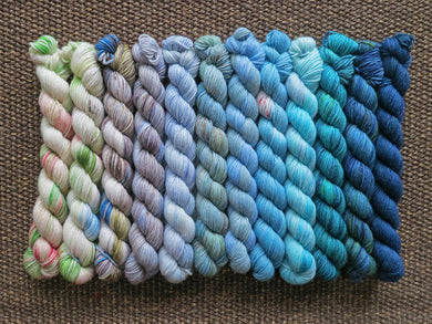 Twelve mini skeins of yarn in various colours ranging from whites and greys through blues and blue greens lined up on a brown woven background