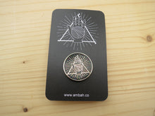 Load image into Gallery viewer, A black glitter enamel pin with Ambah O'Brien's Knitterati logo on a black card with Ambah O'Brien's Knitterati logo, arranged on a pale wooden background