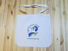 Load image into Gallery viewer, A close up of a natural cotton shoulder tote bag on a yellowed wooden background. The tote bag has a central logo consisting of a multi-coloured ball of yarn held in a white hand. The logo is surrounded by black lettering which reads Dyed By Hand Yarns www.dyedbyhandyarns.com.
