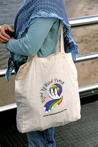 A natural cotton shoulder tote bag with a central logo consisting of a multi-coloured ball of yarn held in a white hand. The logo is surrounded by black lettering which reads Dyed By Hand Yarns www.dyedbyhandyarns.com. The tote bag is on the shoulder of a person wearing a pale blue green long sleeve shirt with a grey blue and white lace shawl and a pair of jeans. In the background are water, sand, railings and black paving.