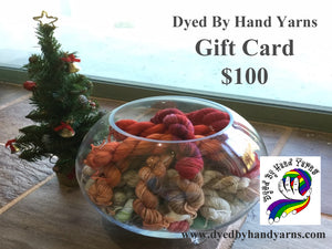 In the foreground is a round glass bowl full of multi-coloured mini skeins; behind this to the left is a decorated mini Christmas tree. This stands on a tan brown stone tile floor. There is dark grey writing in the top right hand corner that says Dyed By Hand Yarns Gift Card $100. In the bottom right hand corner is a multi-coloured logo. Under this in dark grey is a web address