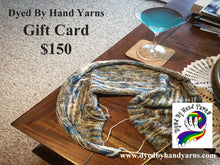 Load image into Gallery viewer, A white, brown and blue variegated piece of knitting on a short circular needle lies on a dark brown coffee table. Also on the coffee table there is a martini glass filled with light blue liquid. In the background is a cream tan flecked carpet. There is dark grey writing in the top left hand corner that says Dyed By Hand Yarns Gift Card $150. In the bottom right hand corner is a multi-coloured logo. Under this in white is a web address
