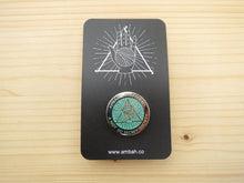 Load image into Gallery viewer, A green glitter enamel pin with Ambah O'Brien's Knitterati logo on a black card with Ambah O'Brien's Knitterati logo, arranged on a pale wooden background