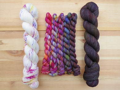 Five brightly coloured variegated mini skeins of yarn flanked by two full sized skeins lined up vertically on a pale wooden background
