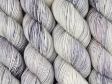 Load image into Gallery viewer, A close up of white and soft grey with speckles of black and purple coloured skeins of superwash bluefaced leicester, silk and cashmere 4ply fingering sock yarn (Newsprint on Blue Chip Stocking)