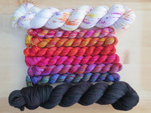 Load image into Gallery viewer, Six brightly coloured variegated mini skeins of yarn flanked by two full sized skeins lined up horizontally on a pale wooden background
