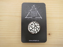 Load image into Gallery viewer, A white glitter enamel pin with Ambah O'Brien's logo on a black card with Ambah O'Brien's Knitterati logo, arranged on a pale wooden background