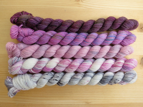 Five variegated mini skeins of yarn in shades of dusty pinks and purples lined up horizontally on a pale wooden background