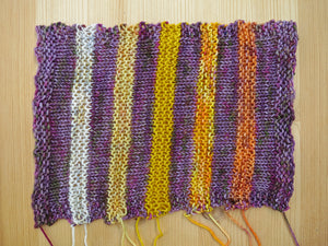 A knitted sample showing orange, yellow and cream stripes alternating with a variegated dusty purple on a pale wooden background