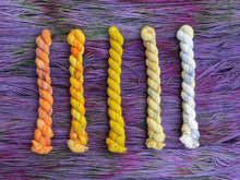 Load image into Gallery viewer, Five variegated mini skeins of yarn in shades of orange, yellows and cream lined up vertically on a background of variegated dusty purple yarn 
