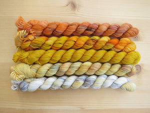 Five variegated mini skeins of yarn in shades of orange, yellows and cream lined up horizontally on a pale wooden background