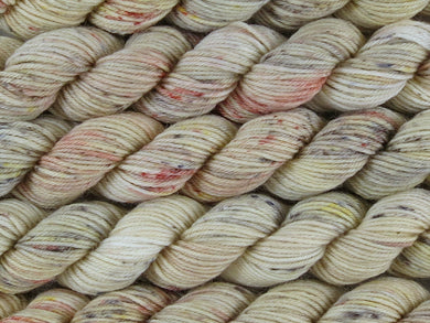 A close up of variegated ecru with speckles of rust, gold, brown and charcoal coloured mini skeins of superwash merino and nylon 4ply fingering sock yarn arranged horizontally (Liquor Is Quicker on Tough Stocking Mini)
