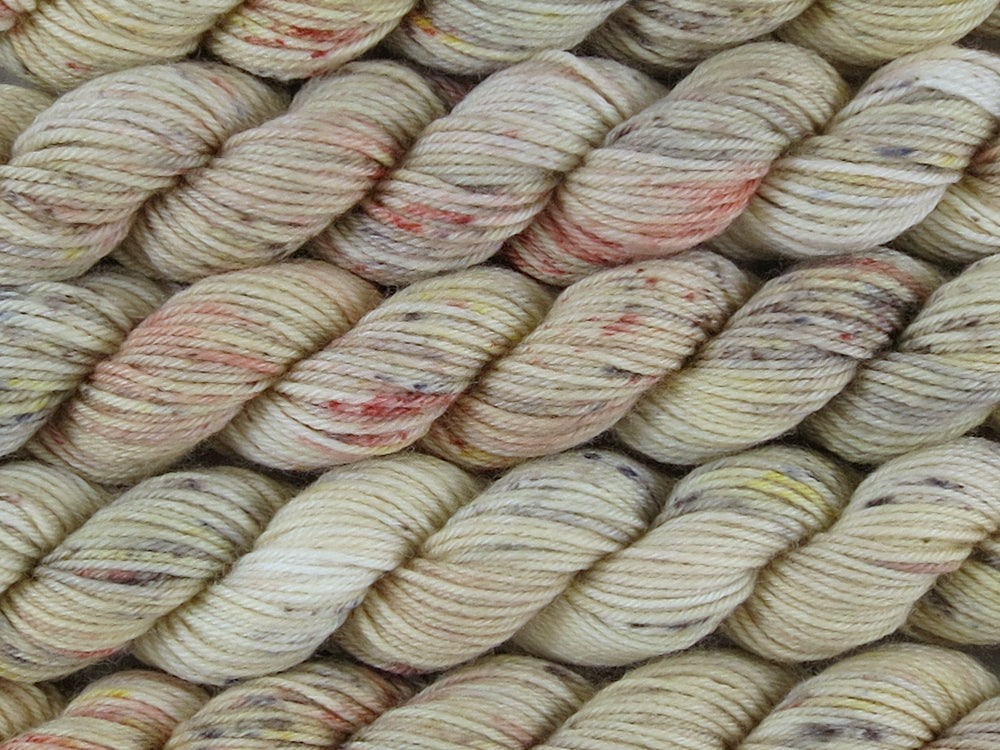 A close up of variegated ecru with speckles of rust, gold, brown and charcoal coloured mini skeins of superwash merino and nylon 4ply fingering sock yarn arranged horizontally (Liquor Is Quicker on Tough Stocking Mini)