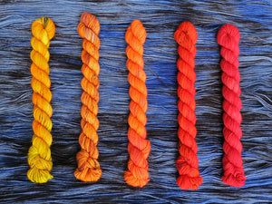 Five variegated mini skeins of yarn in shades of red, orange and yellows lined up vertically on a background of variegated blue and black yarn 