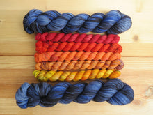 Load image into Gallery viewer, Five variegated mini skeins of yarn in shades of red, orange and yellows flanked by two full sized variegated blue and black lined up horizontally on a pale wooden background
