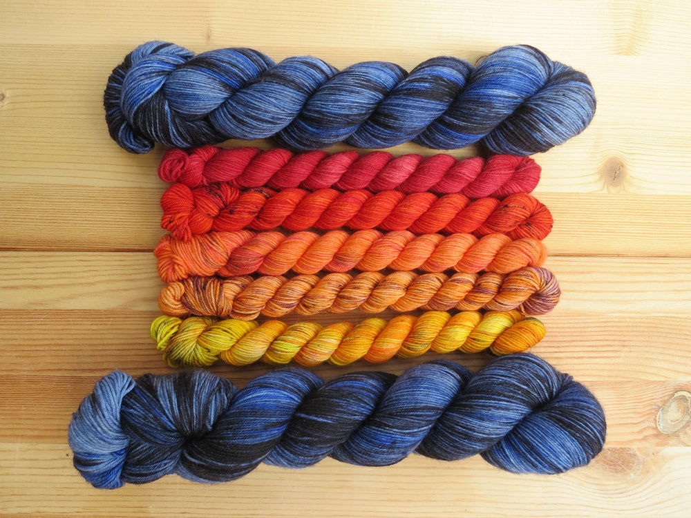 Five variegated mini skeins of yarn in shades of red, orange and yellows flanked by two full sized variegated blue and black lined up horizontally on a pale wooden background