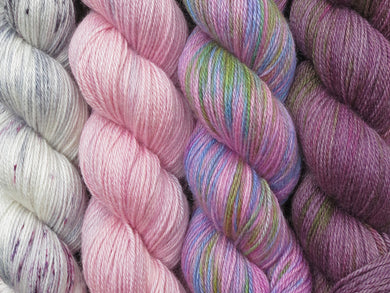 A close up of four skeins of yarn in various shades of pink and purple from variegated white with grey and pink on the left through to a variegated purple and green on the right (Solaris Geogradient Yarn Kit on Blue Chip Stocking)