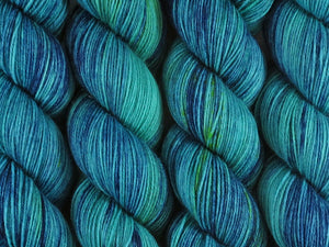 A close up of variegated teal greens and navy blue with speckles of bright yellow coloured skeins of superwash merino and nylon 4ply fingering sock yarn (The Other Woman on Tough Stocking)