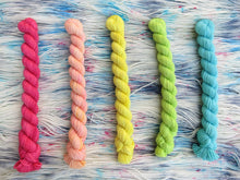 Load image into Gallery viewer, Five light bright rainbow coloured mini skeins of yarn lined up vertically on a background of variegated white, sky blue and grey with pink speckles yarn 
