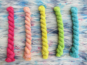Five light bright rainbow coloured mini skeins of yarn lined up vertically on a background of variegated white, sky blue and grey with pink speckles yarn 