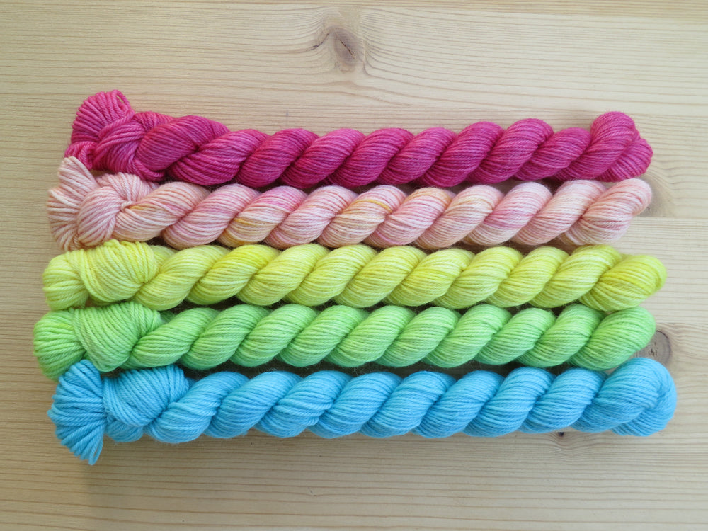 Five light bright rainbow coloured mini skeins of yarn lined up horizontally on a pale wooden background