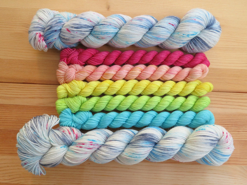 Five light bright rainbow coloured mini skeins of yarn flanked by two full sized variegated white, sky blue and grey with pink speckles skeins lined up horizontally on a pale wooden background