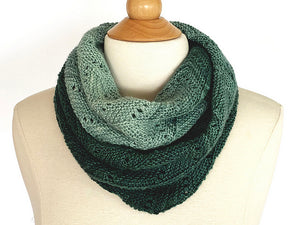 A graduated coloured lace knitted cowl in shades of green double looped on the neck of a cream dress mannequin on a white background. Pattern is Adventurer Cowl by Ambah O'Brien