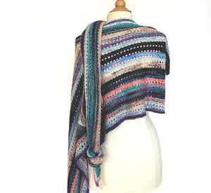 A striped knitted wrap made from twenty five mini skeins arranged in random colour order of purples, greens, whites and pinks draped over a white dress mannequin on a white background. One end of the wrap is knotted and hangs low over the shoulder (Adventuring Wrap by Ambah O'Brien)