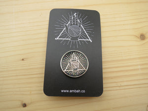 A black glitter enamel pin with Ambah O'Brien's Knitterati logo on a black card with Ambah O'Brien's Knitterati logo, arranged on a pale wooden background