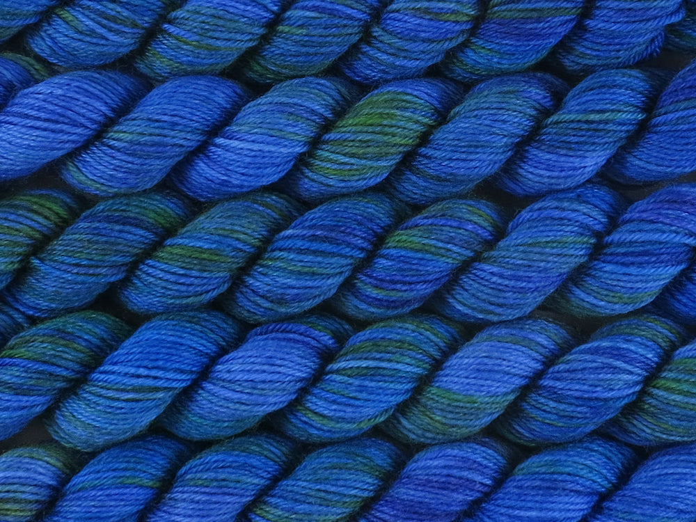 A close up of variegated bright blue, deep violet and emerald green mini skeins of superwash merino and nylon 4ply fingering sock yarn arranged horizontally (Blue Quandong on Tough Stocking Mini)