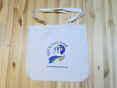 A close up of a natural cotton shoulder tote bag on a yellowed wooden background. The tote bag has a central logo consisting of a multi-coloured ball of yarn held in a white hand. The logo is surrounded by black lettering which reads Dyed By Hand Yarns www.dyedbyhandyarns.com.