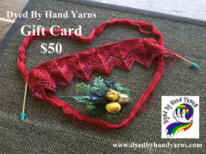 Red lace knitting on a short circular needle sits on a red skein of yarn arranged in the shape of a heart, all on top of a textured brown jute rug. There is white writing in the top left hand corner that says Dyed By Hand Yarns Gift Card $50. In the bottom right hand corner is a multi-coloured logo. Under this in white is a web address