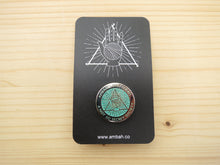 Load image into Gallery viewer, A green glitter enamel pin with Ambah O&#39;Brien&#39;s Knitterati logo on a black card with Ambah O&#39;Brien&#39;s Knitterati logo, arranged on a pale wooden background
