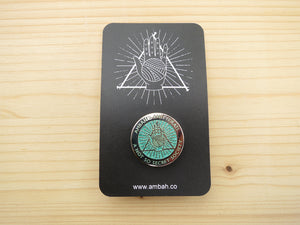 A green glitter enamel pin with Ambah O'Brien's Knitterati logo on a black card with Ambah O'Brien's Knitterati logo, arranged on a pale wooden background