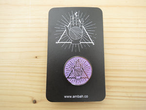 A mauve glitter enamel pin with Ambah O'Brien's Knitterati logo on a black card with Ambah O'Brien's Knitterati logo, arranged on a pale wooden background