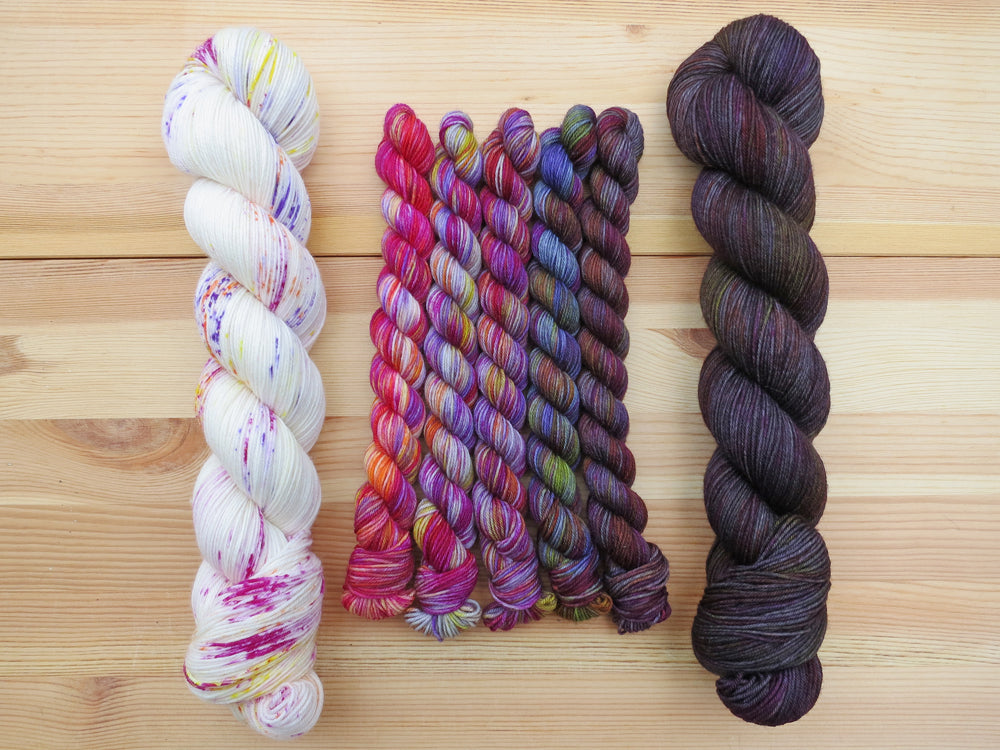 Five brightly coloured variegated mini skeins of yarn flanked by two full sized skeins lined up vertically on a pale wooden background