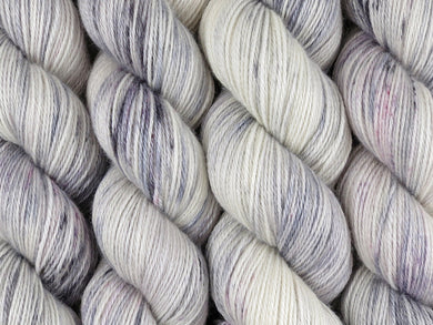 A close up of white and soft grey with speckles of black and purple coloured skeins of superwash bluefaced leicester, silk and cashmere 4ply fingering sock yarn (Newsprint on Blue Chip Stocking)