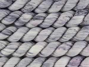 A close up of white and soft grey with speckles of black and purple mini skeins of superwash merino and nylon 4ply fingering sock yarn arranged horizontally (Newsprint on Tough Stocking Mini)